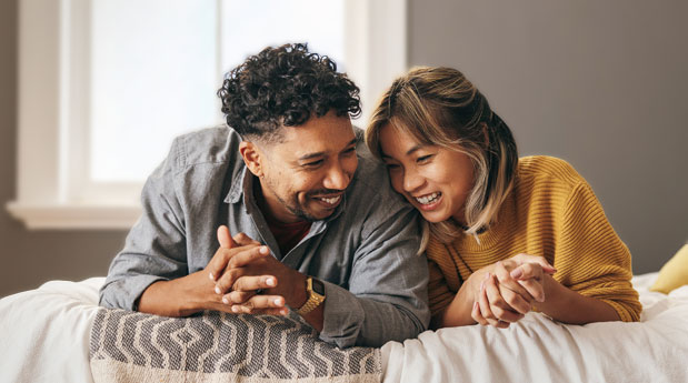 A couple lays across a bed, propped up on their elbows. They are leaning into each other and smiling.