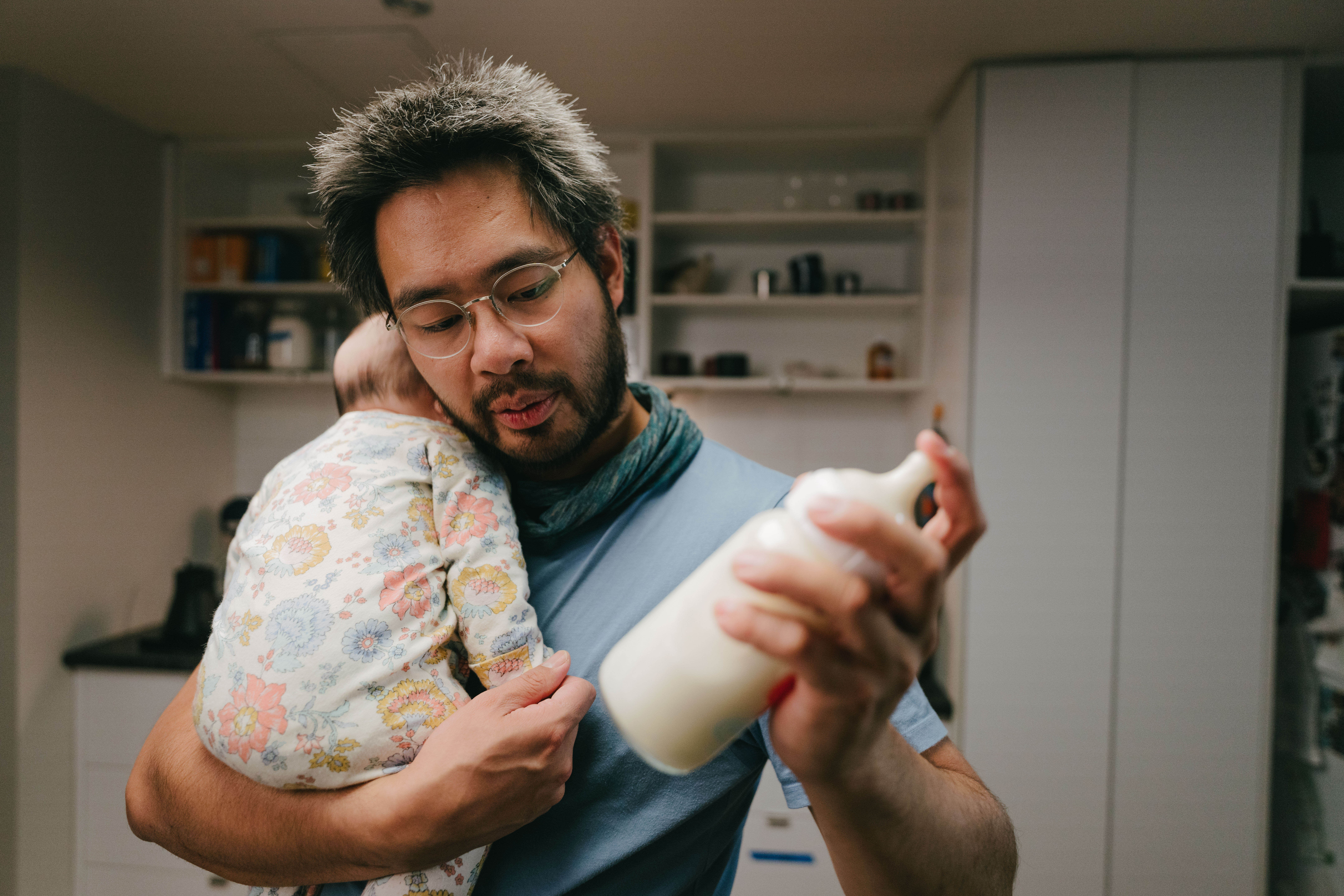 A dad holds his baby in one arm and makes a bottle with his other hand.