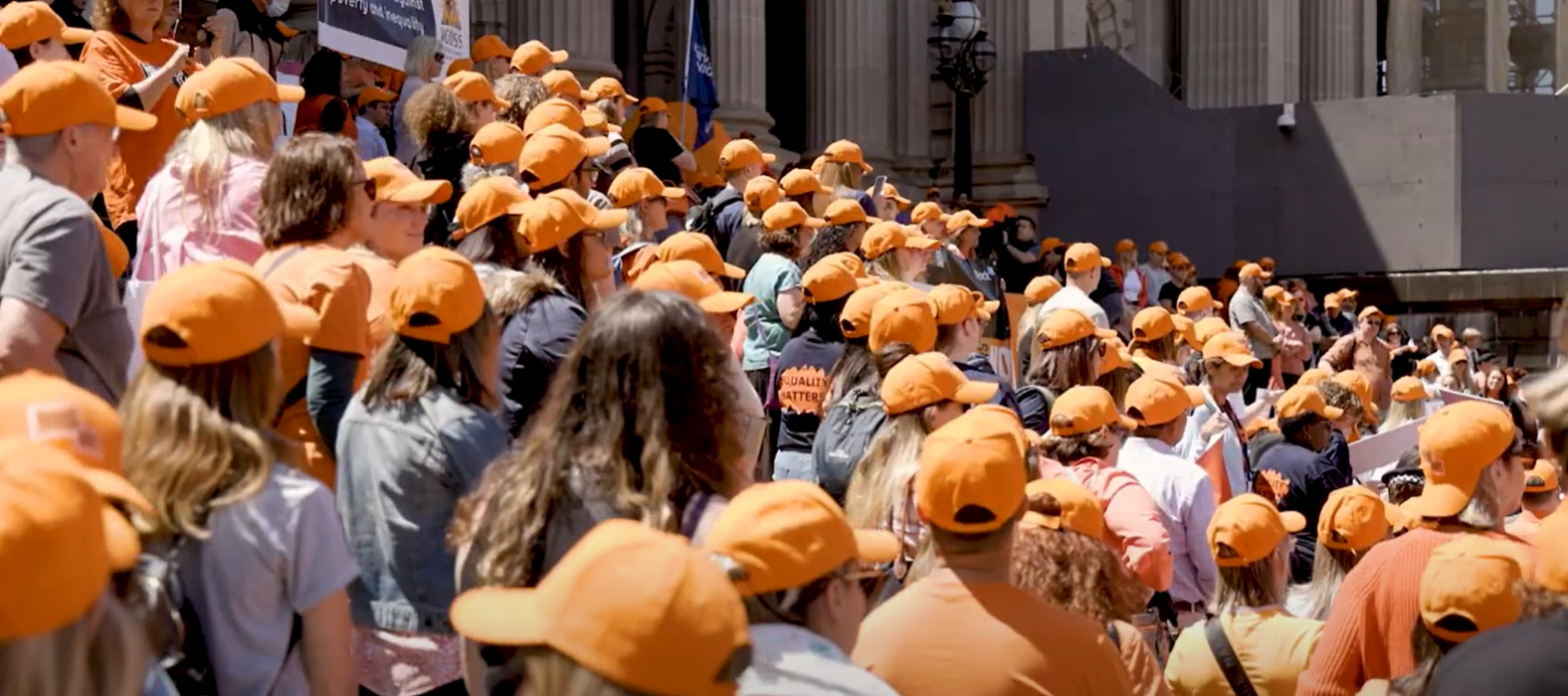 A crowd of people wearing orange caps stand on the steps of Parliament House, Melbourne, listening to speeches at the 2022 Walk Against Family Violence.