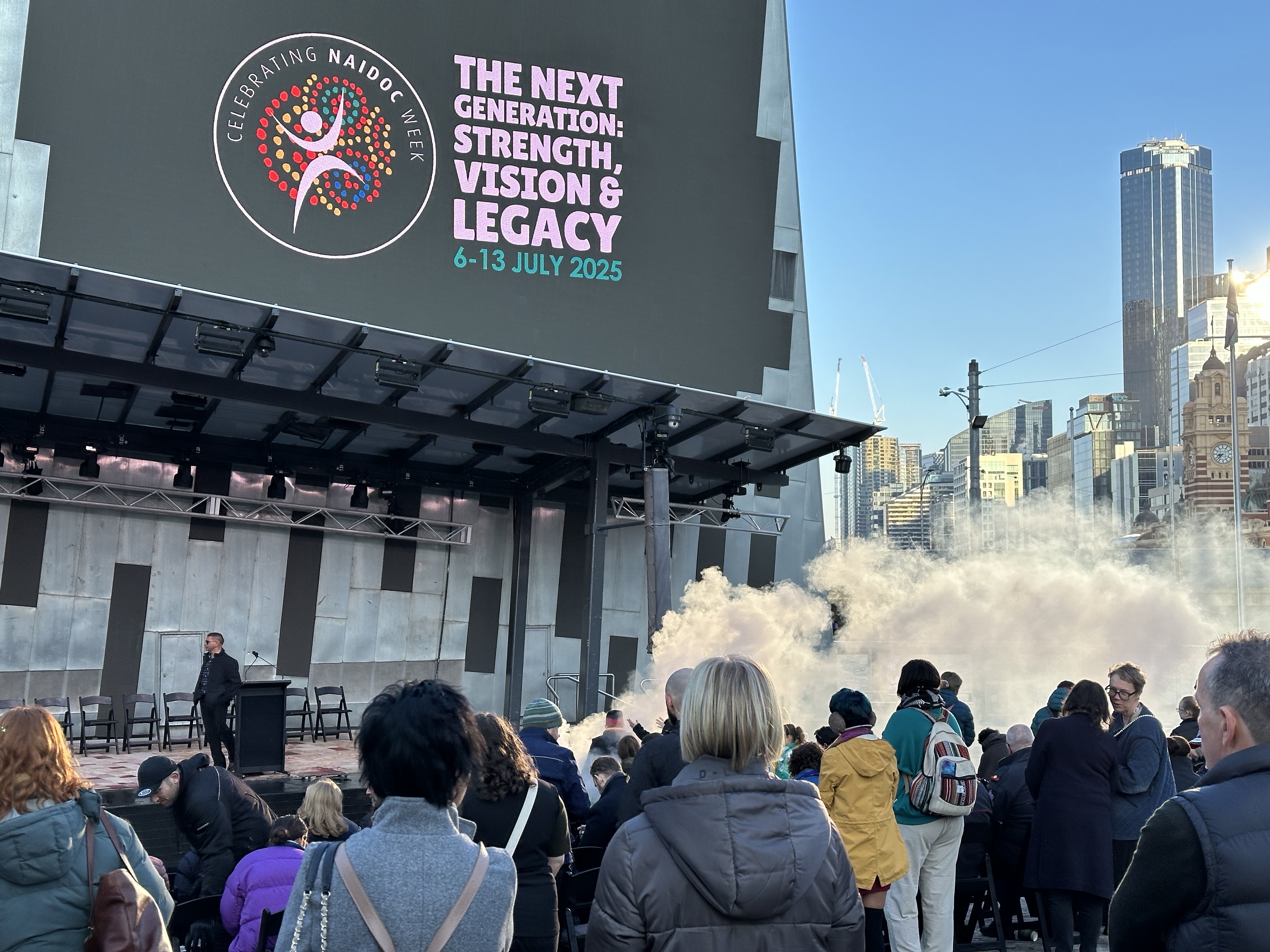 People gather at Federation square for the flag-raising ceremony. Smoke from a smoking ceremony billows out under a large sign that says 'The Next Generation: Strength, Vision and Legacy 6-13 July 2025'