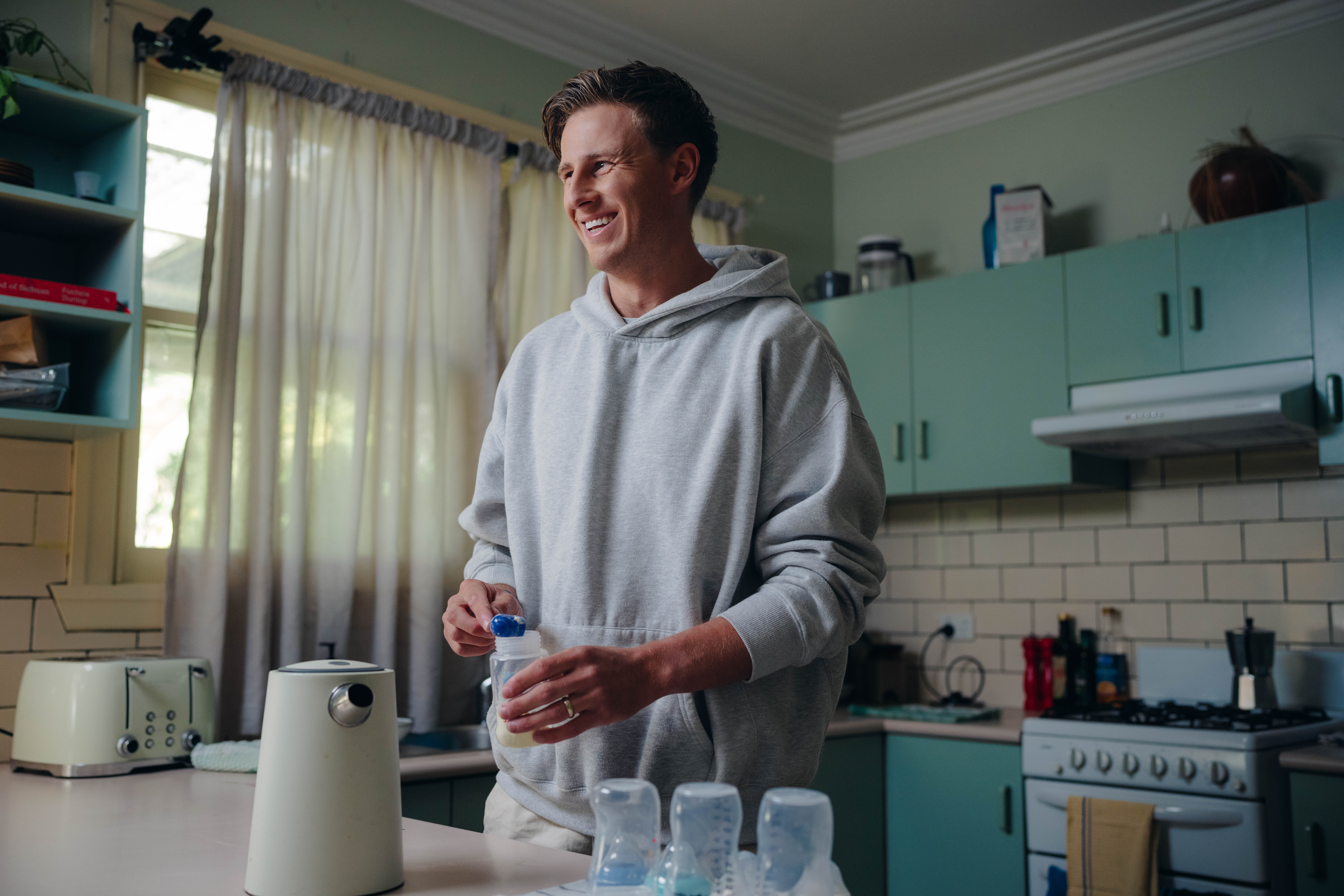 Young father wearing grey hooded jumper, standing at kitchen counter preparing baby formula in bottles