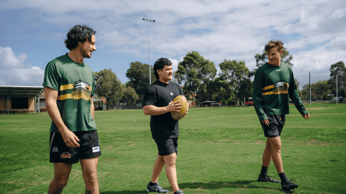 Three men walking together on a community football field, dressed for footy practice, one man holding an Aussie Rules football.