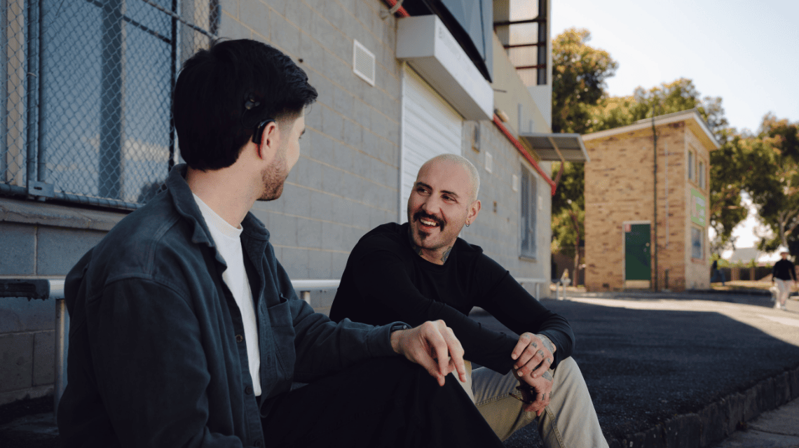 Two men sit on the ground, in casual conversation. 