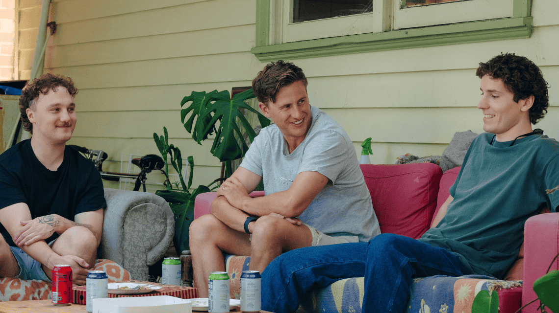 Three men in their 20s sit chatting on a couch on a house porch.