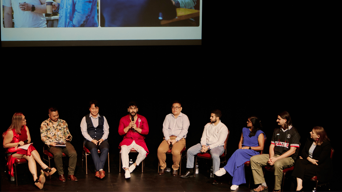 A panel discussion from the launch of the multicultural phase of What Kind of Man Do You Want to Be?. 9 people sit in a row on a stage. 