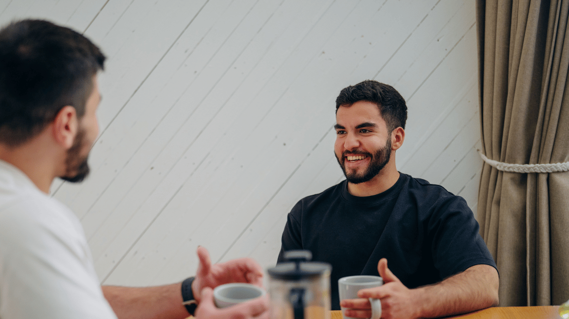 A man in a black T-shirt drinking coffee in happy conversation with a friend.