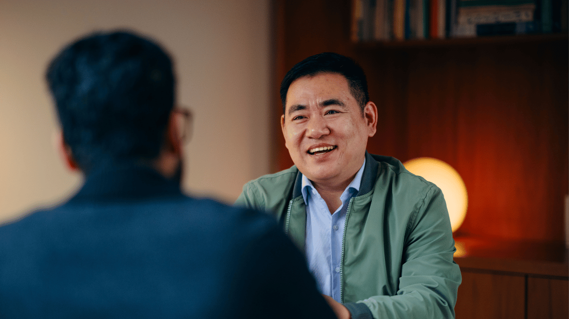 A man in a green jacket is in a home office, sitting in conversation across from a friend.