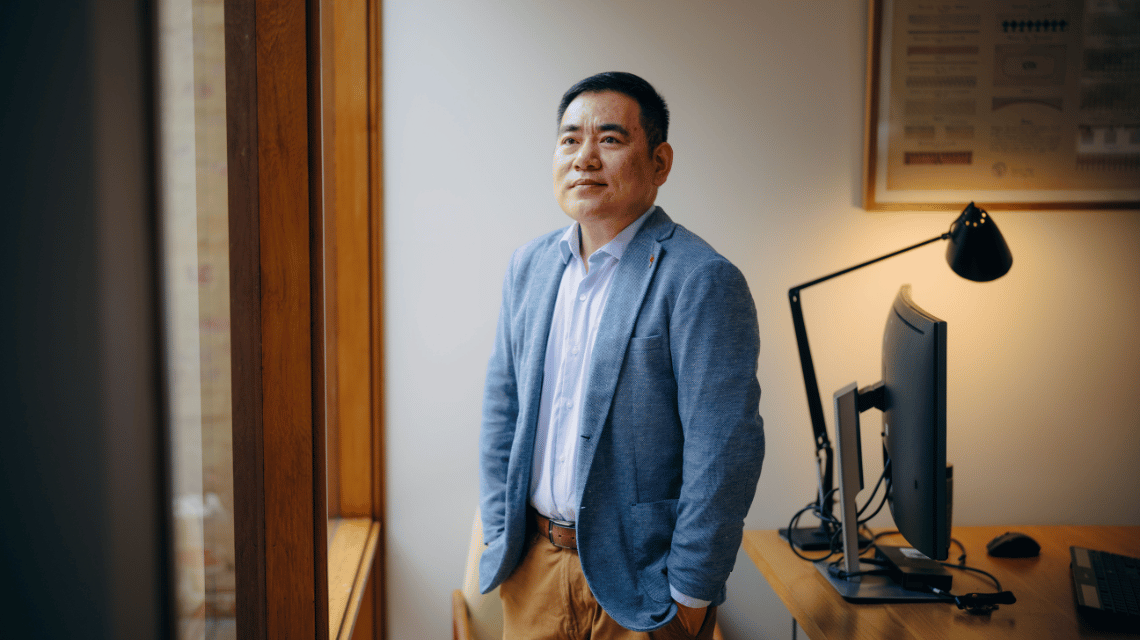 A man in a green jacket stands in front of his home office desk, looking thoughtfully out the window.