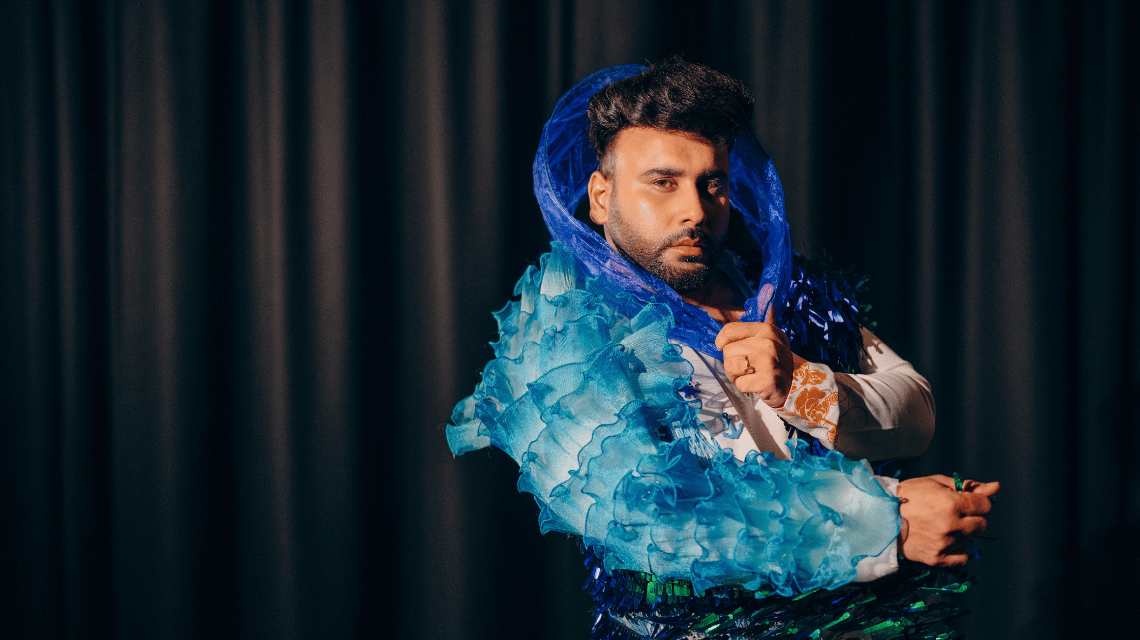 A man poses to showcase his pageant costume; a detailed outfit in shades of blue.