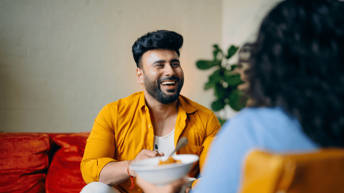 A laughing man shares a casual meal on a couch with a friend.