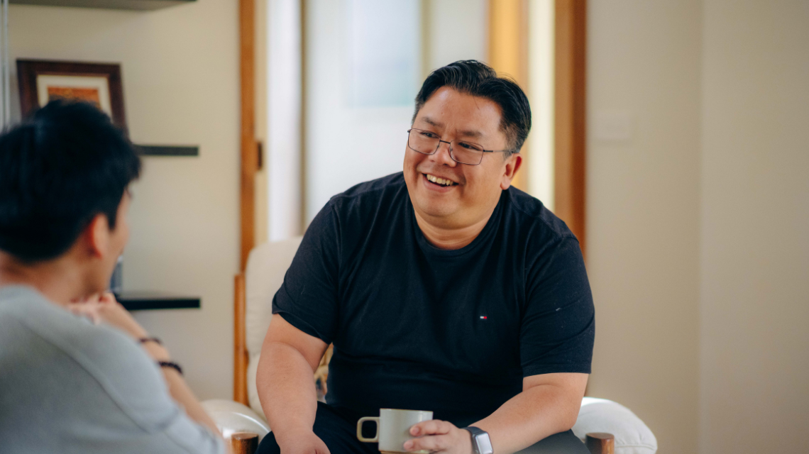 A man in a black t-shirt is sitting in a loungeroom, laughing in conversation with a friend.
