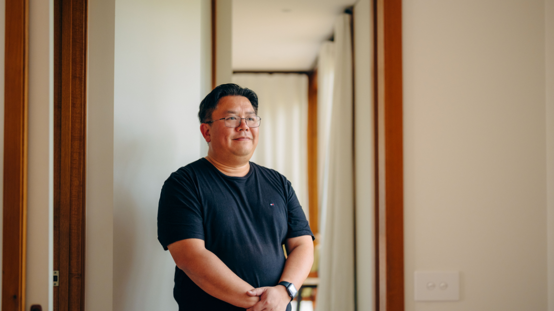 A man in a black t-shirt smiles to the camera, standing in a home hallway.