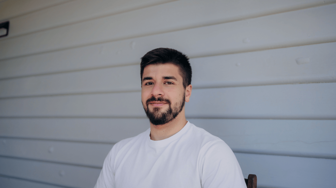 Portrait of a man in a white t-shirt sitting in front of a weatherboard house.