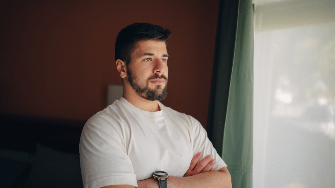 Portrait of a man in a white t-shirt looking out a window.