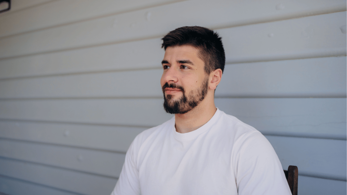 Portrait of a man in a white t-shirt sitting outside a weatherboard house, looking away.