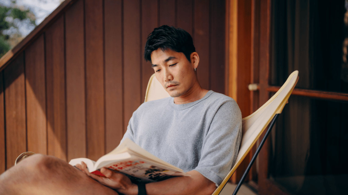 A man in a grey t-shirt relaxes on a home balcony, reading a book. 