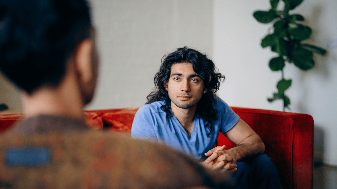 A man with black wavy hair sits on a red couch, leaning forward listening to a friend.