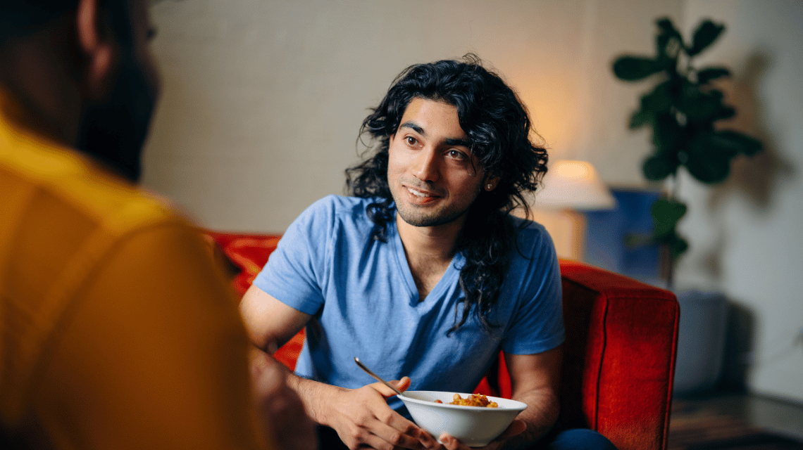 A man with dark wavy hair shares a relaxed meal with a friend.