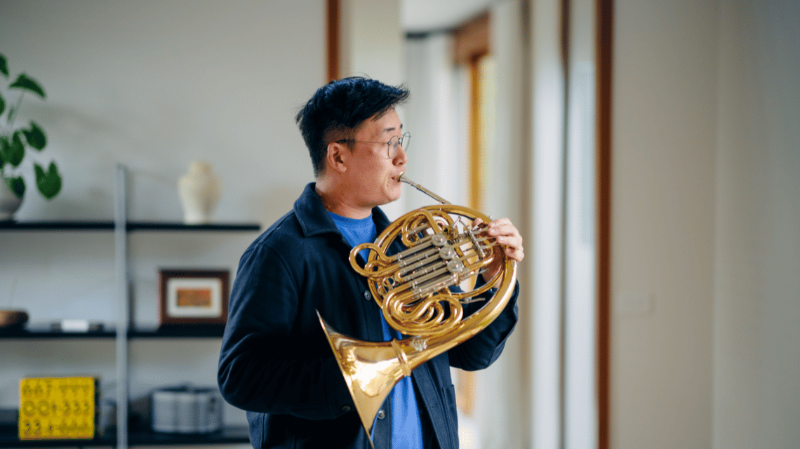 A portrait of a man in a living room, playing a French horn.