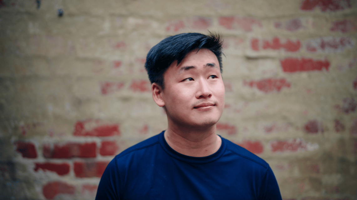 Portrait of a man in a blue sweater, standing in front of a brick wall.