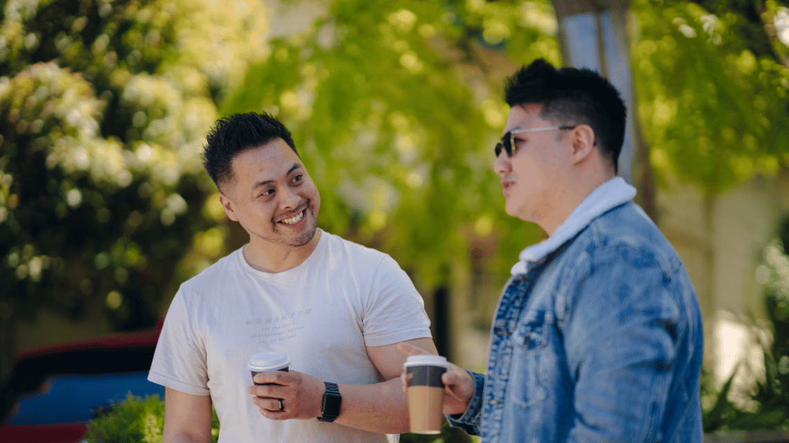 Two men walk in a leafy street, holding takeaway coffee.