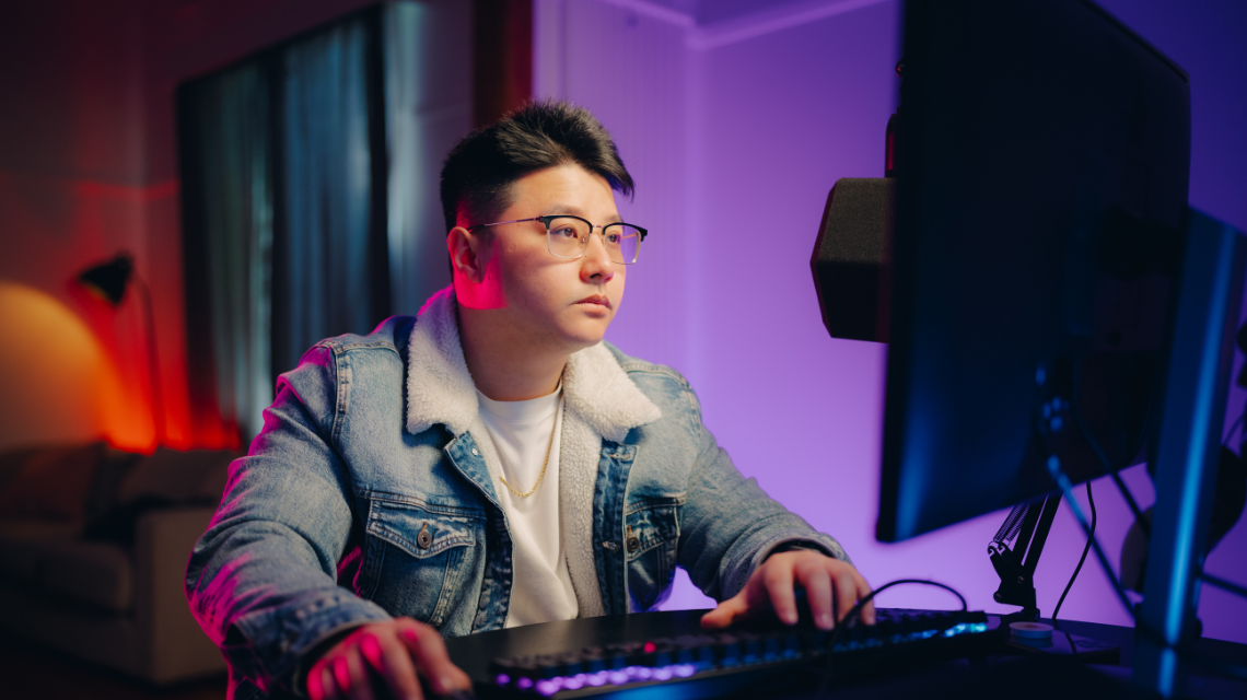 A man in a denim jacket sits at a gaming computer. The room is lit with purple.
