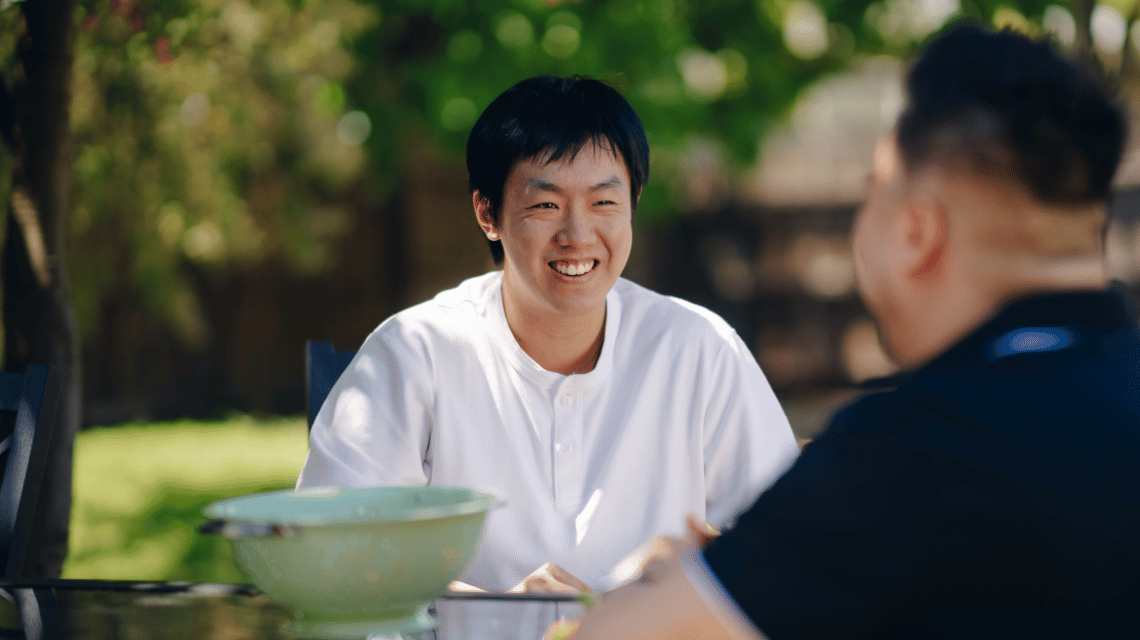 A man in a white t-shirt sits at a backyard table, smiling at his friend sitting opposite.