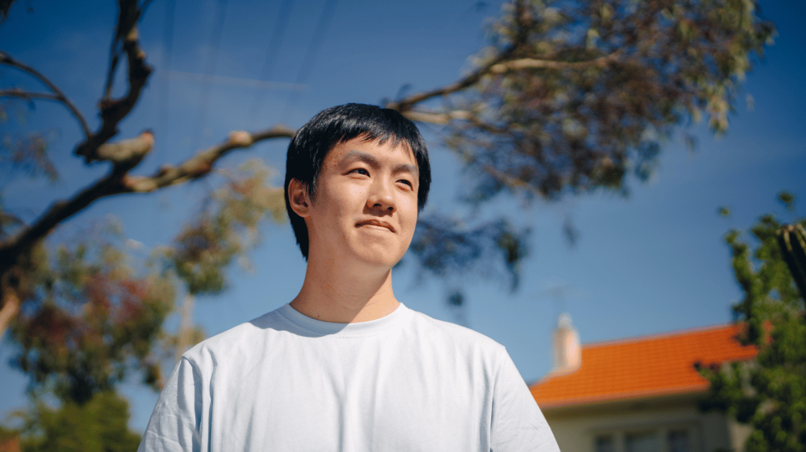 A man in a white t-shirt stands outside a suburban home, under a gum tree.