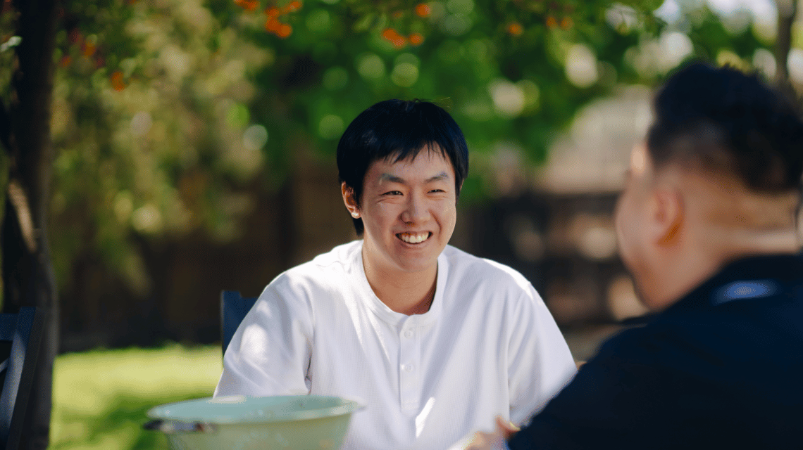 Two men sit at a table in a backyard and chat.