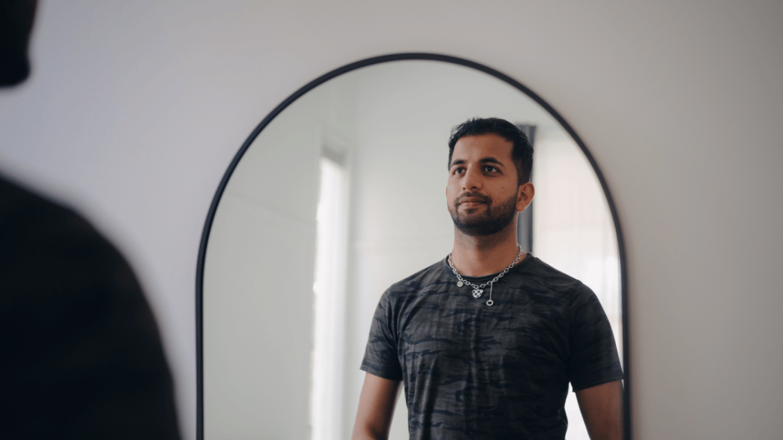 A man in a grey t-shirt checks his reflection in a mirror.
