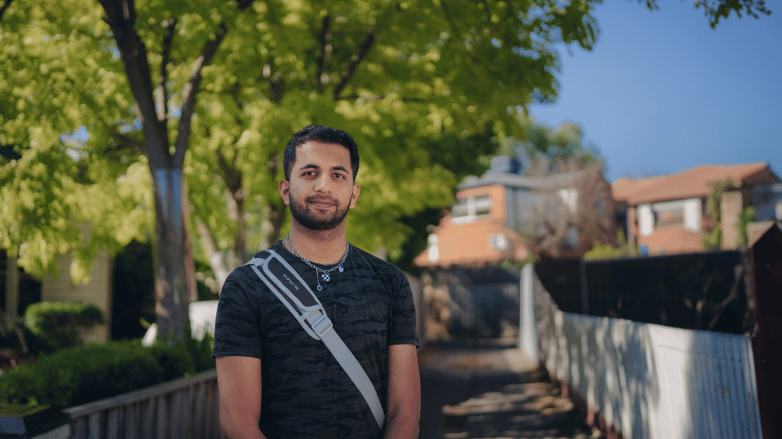 A man in a grey stands in a leafy suburban driveway.