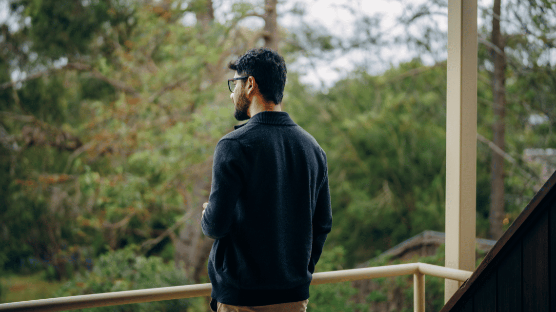 A man looks out at a leafy garden from a balcony.