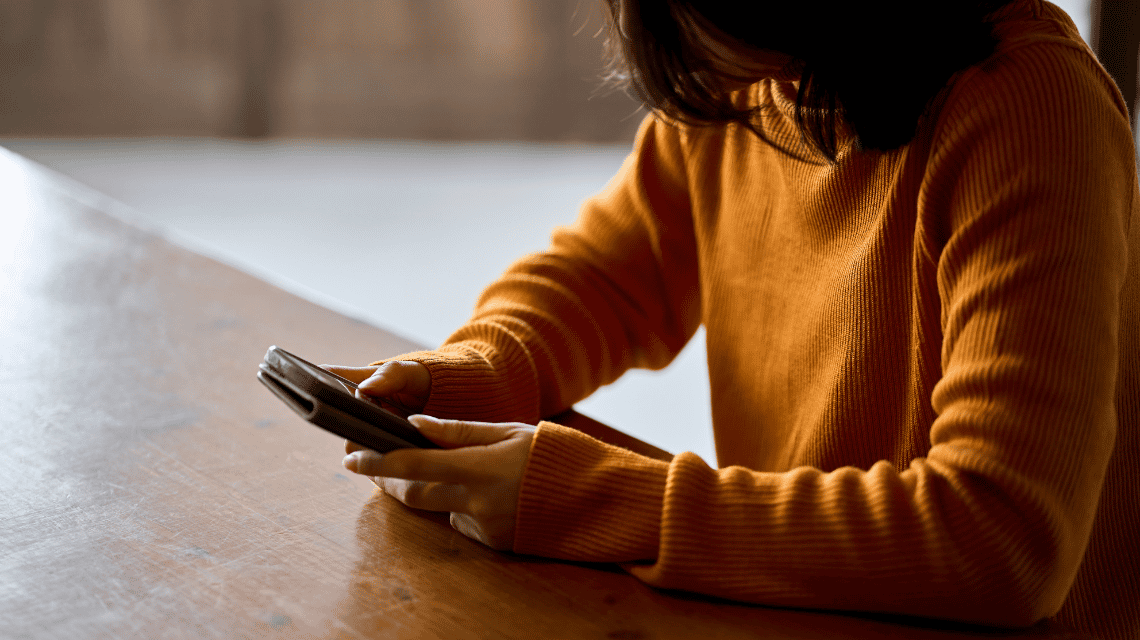 The shoulders and arms of a woman in an orange jumper, sitting at a table checking the messages on her phone. 