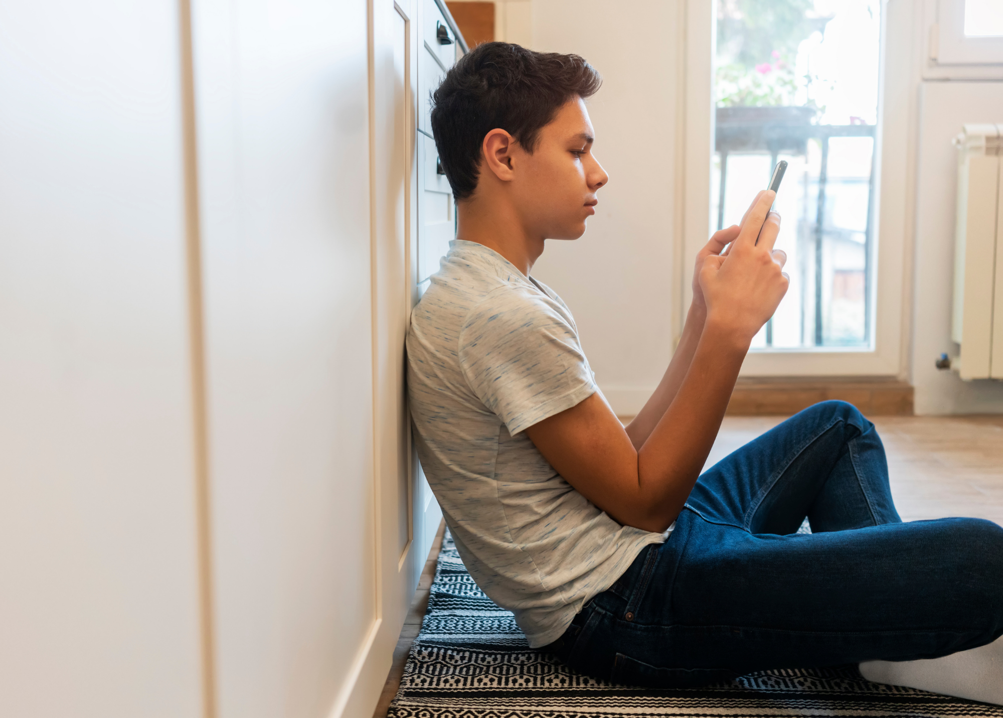 A teenage boy sitting on the floor leaning against a cabinet while looking at his smartphone