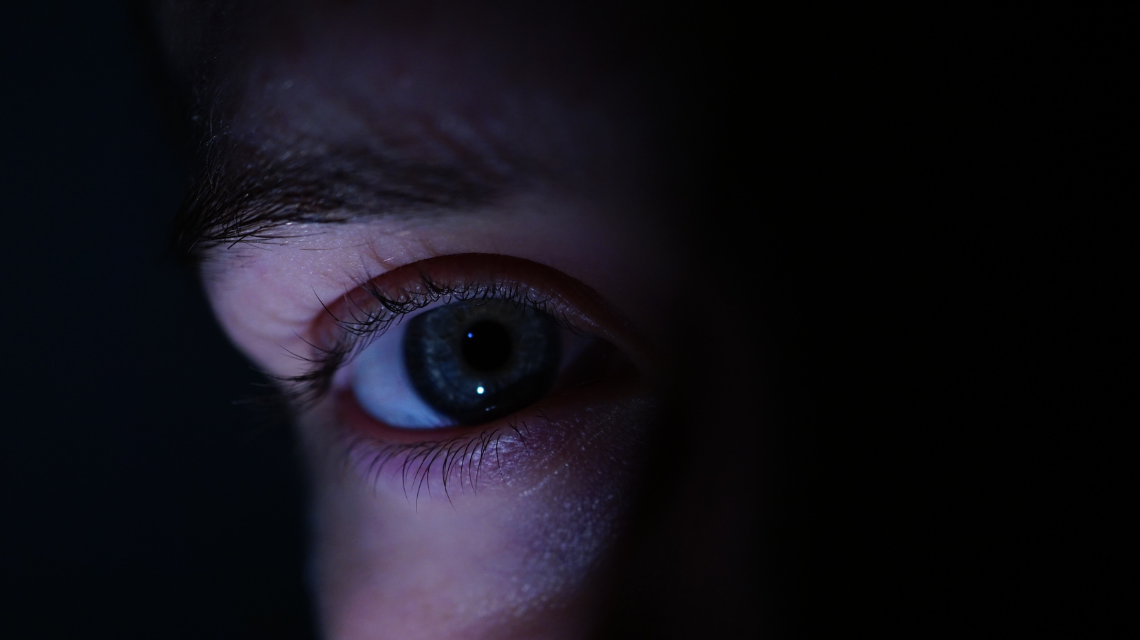 A close-up of a human eye glowing on a smartphone screen in a dark room, symbolising digital surveillance